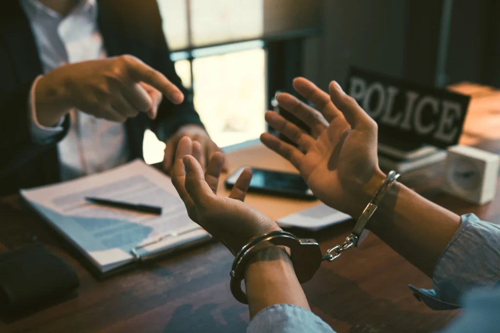 A person in handcuffs sits across from a criminal defense attorney during a legal consultation, with a police sign visible in the background — representing felony defense in Tacoma.