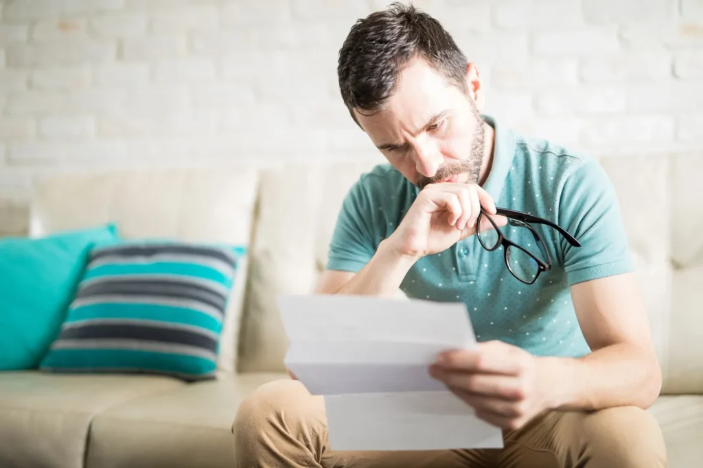 Concerned man sitting on couch reading official documents, symbolizing stress or confusion about a legal notice or court order.